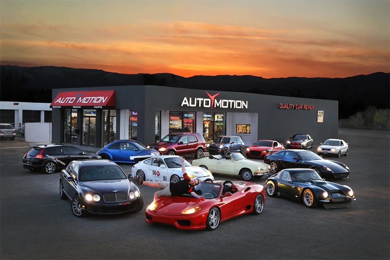 Twilight exterior of the AutoMotion quality car repair/dealership building with an arranged fleet of luxury and sports cars (red Ferrari convertible, Bentley, Porsches) in the lot; orange sunset sky over dark hills, lit signage.