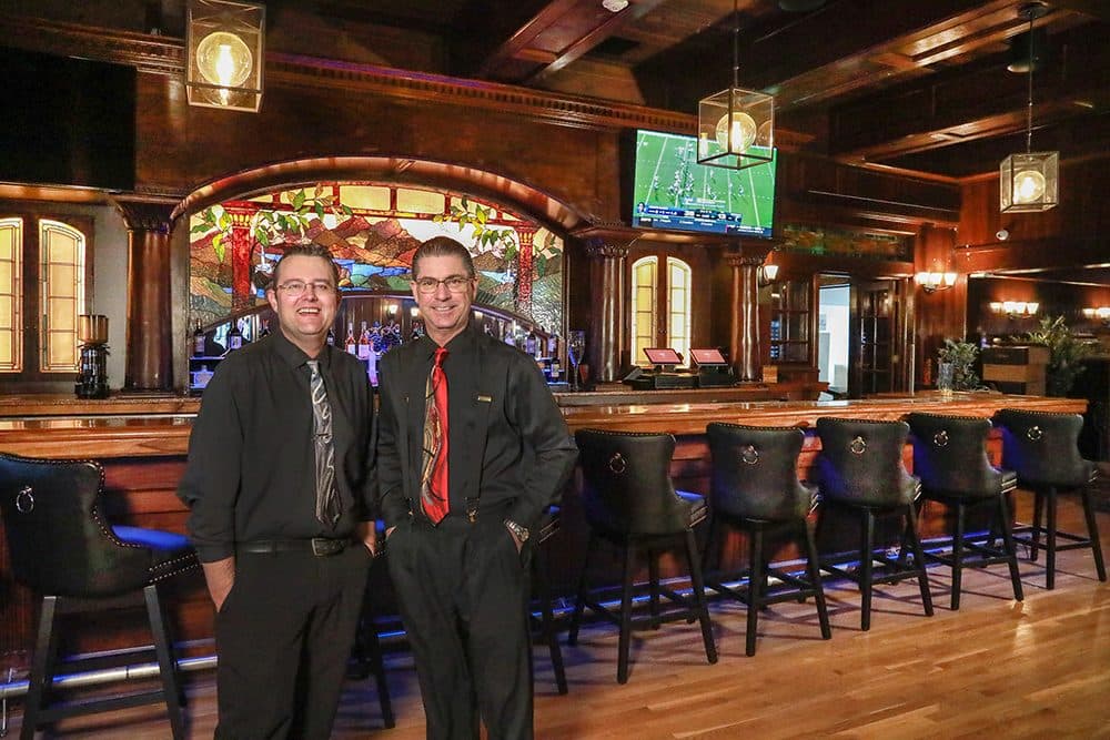 Two Marcolino's managers in suits stand in the renovated dark-wood bar, warm tungsten light, stained glass behind