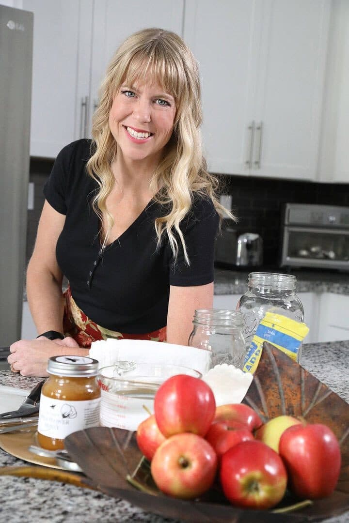 Nutritionist Carly Neubert smiling in a white kitchen behind a bowl of red apples and vinegar ingredients, soft daylight