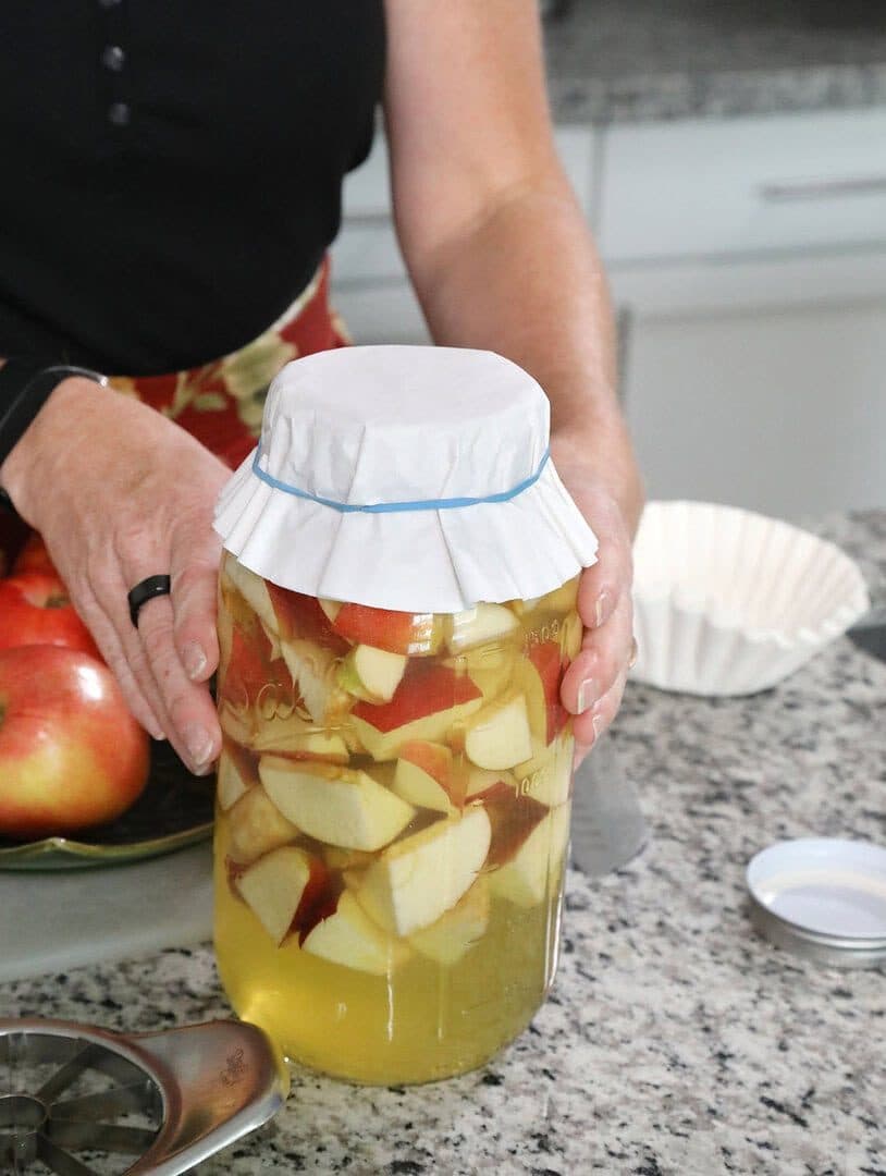 Hands covering a jar of chopped apples in honey-water with a coffee filter, granite counter, natural light
