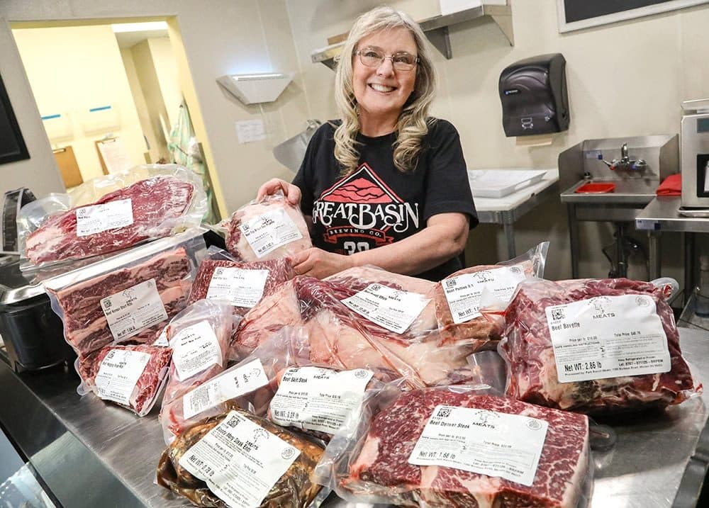 Butcher-shop manager smiling over a counter piled with packaged beef cuts, bright fluorescent kitchen