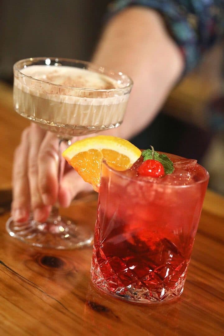 Hand presenting a creamy coupe cocktail and a red cocktail with orange wheel on a wood bar, dark warm background