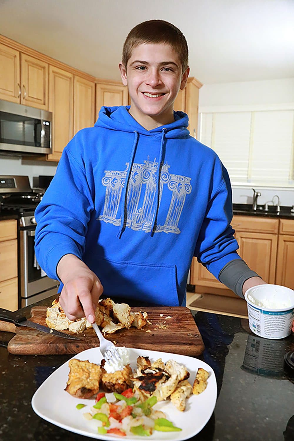 Teen athlete in a blue hoodie plating grilled chicken and vegetables in a home kitchen