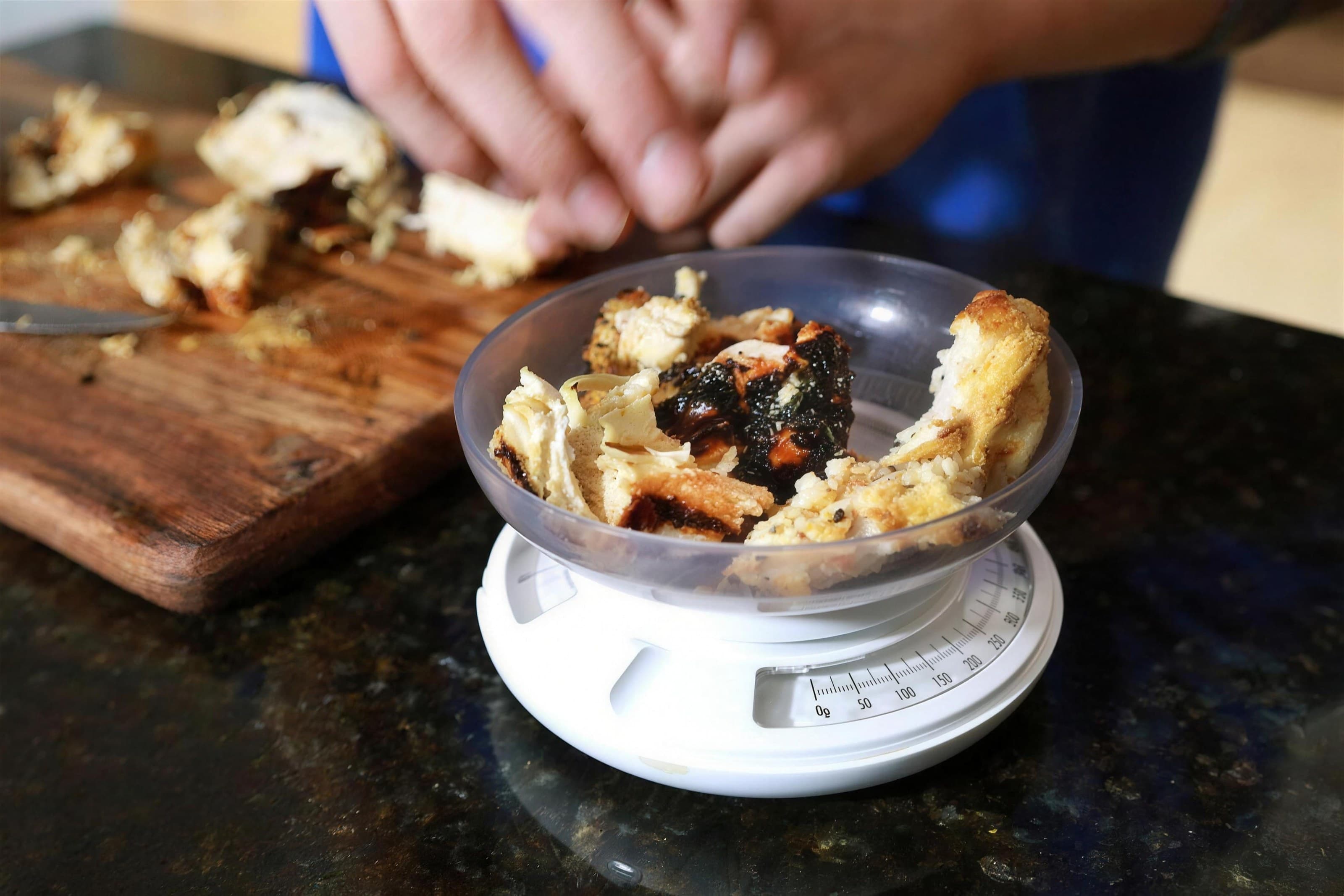 Grilled chicken pieces in a bowl on a kitchen scale beside a cutting board, dark granite counter