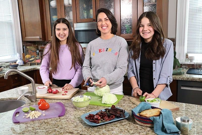 Mother and two daughters smiling while prepping pozole ingredients at a kitchen island
