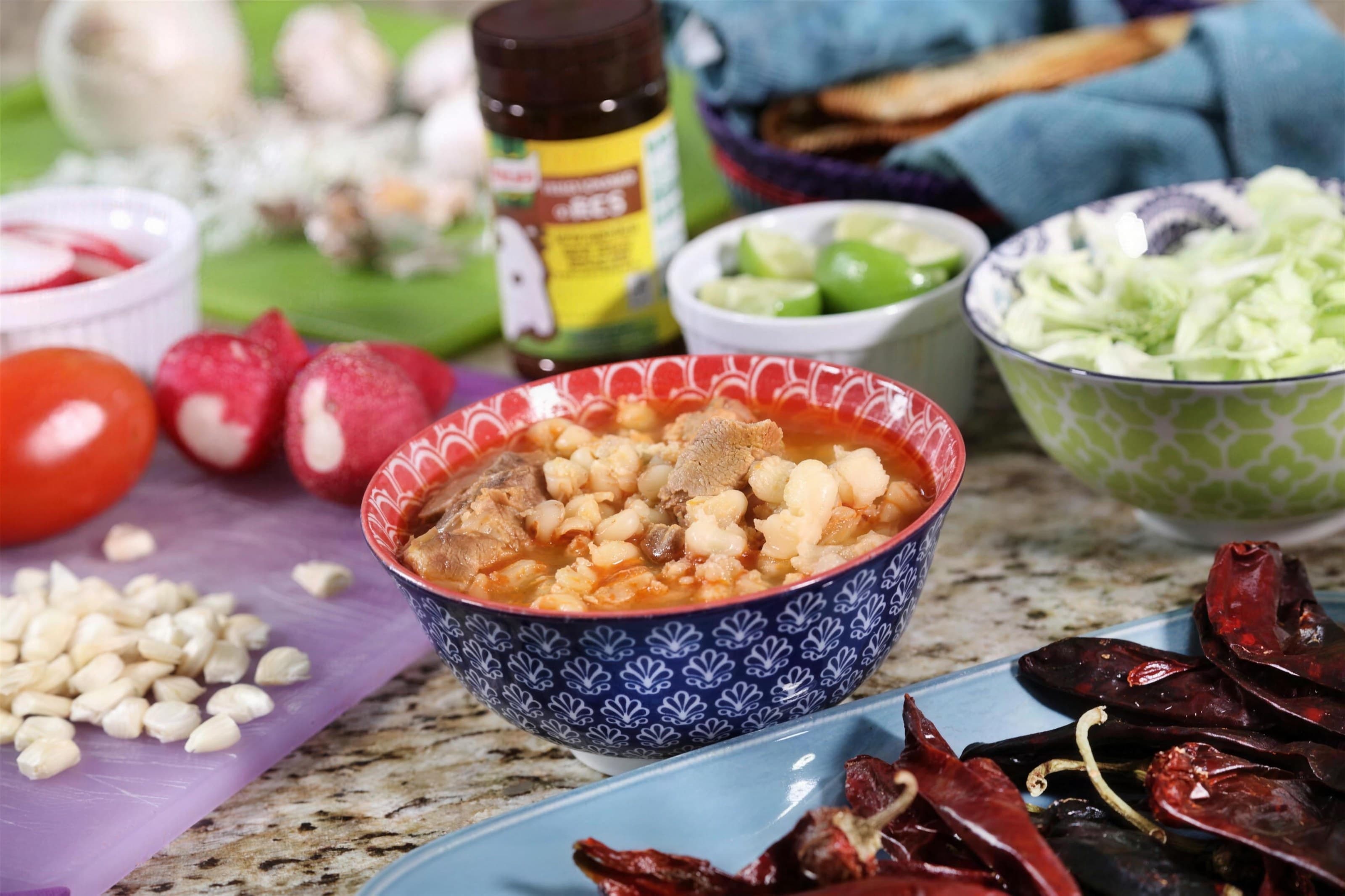 Blue patterned bowl of pozole surrounded by dried chiles, radishes, cabbage and garnishes, bright styled table