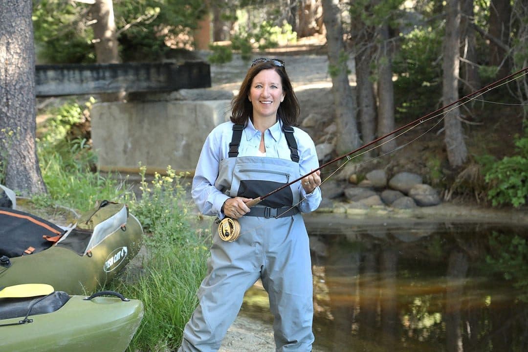 Wildlife commissioner in waders holding a fly rod beside a pond, forest backdrop, natural light