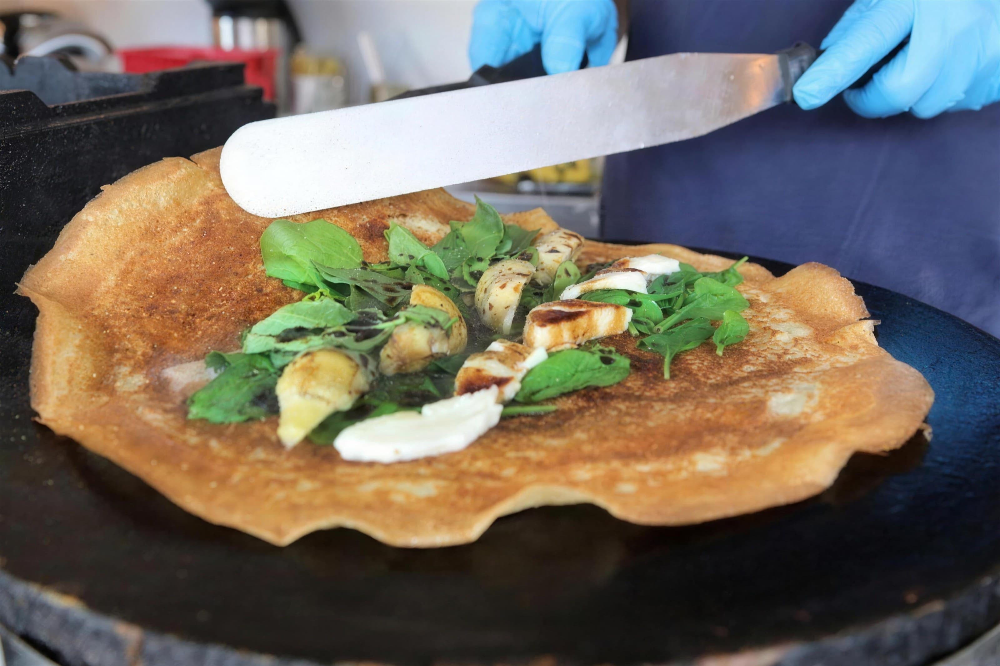 Savory crêpe on the griddle topped with greens, chicken and drizzle, spatula overhead, dark background