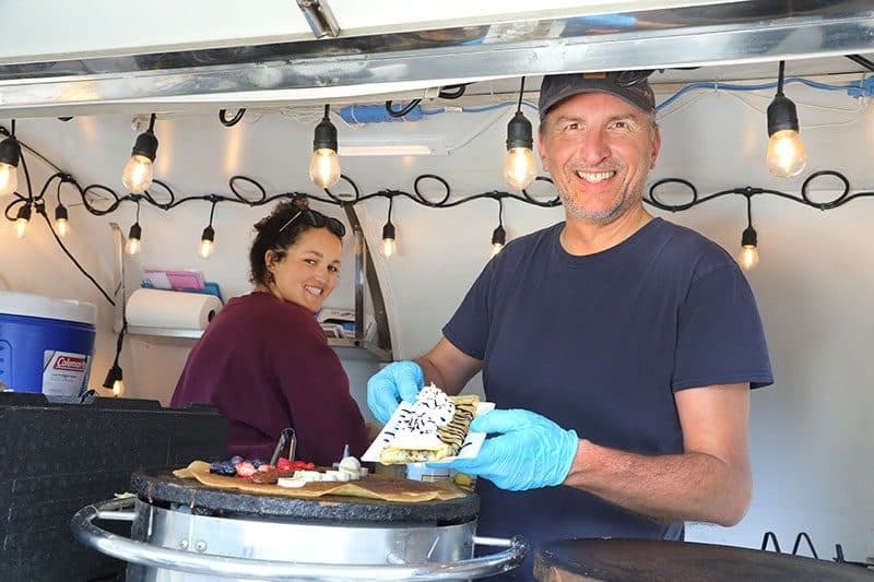 Crêpe maker in a cap smiling in his food trailer with string lights, holding a folded crêpe