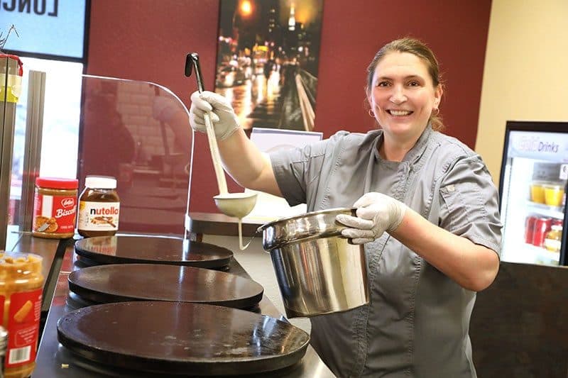 Kaffé Crepe owner in chef coat ladling batter onto a round crêpe griddle, red-walled café