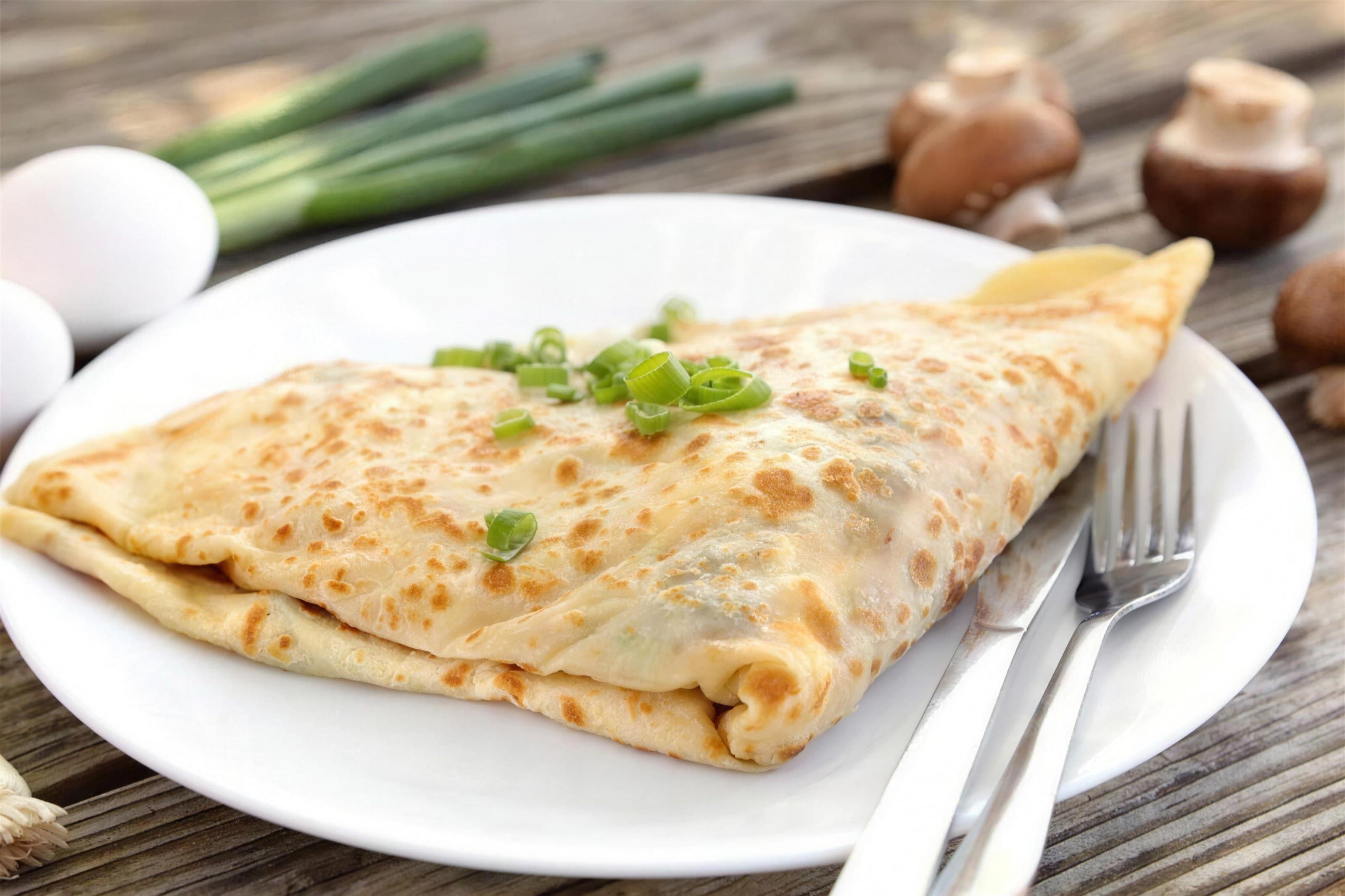 Folded breakfast crêpe with scallions on a white plate, mushrooms and eggs on rustic wood, soft daylight