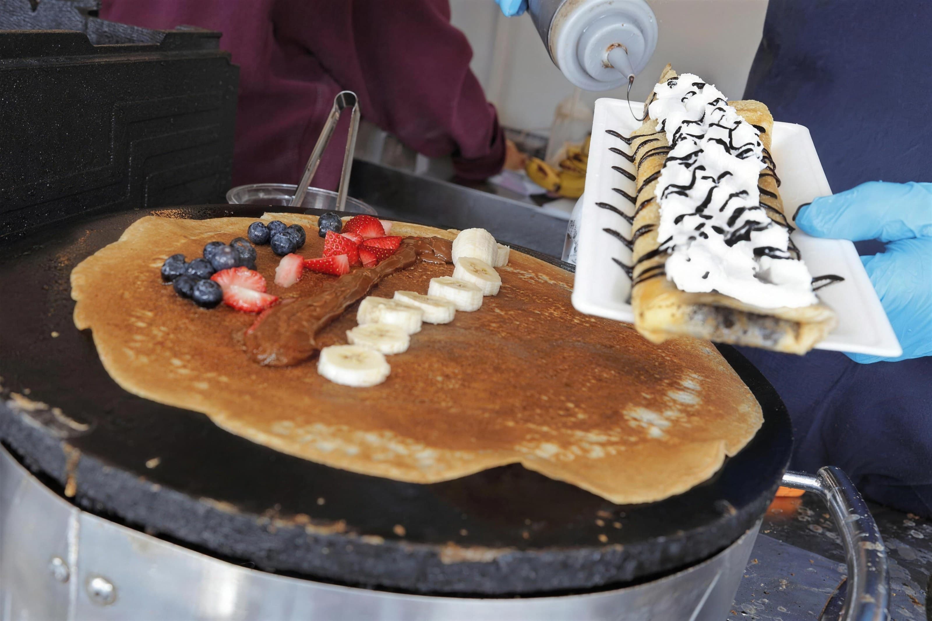 Banana, strawberry and blueberry crêpe on the griddle beside a plated crêpe with chocolate drizzle