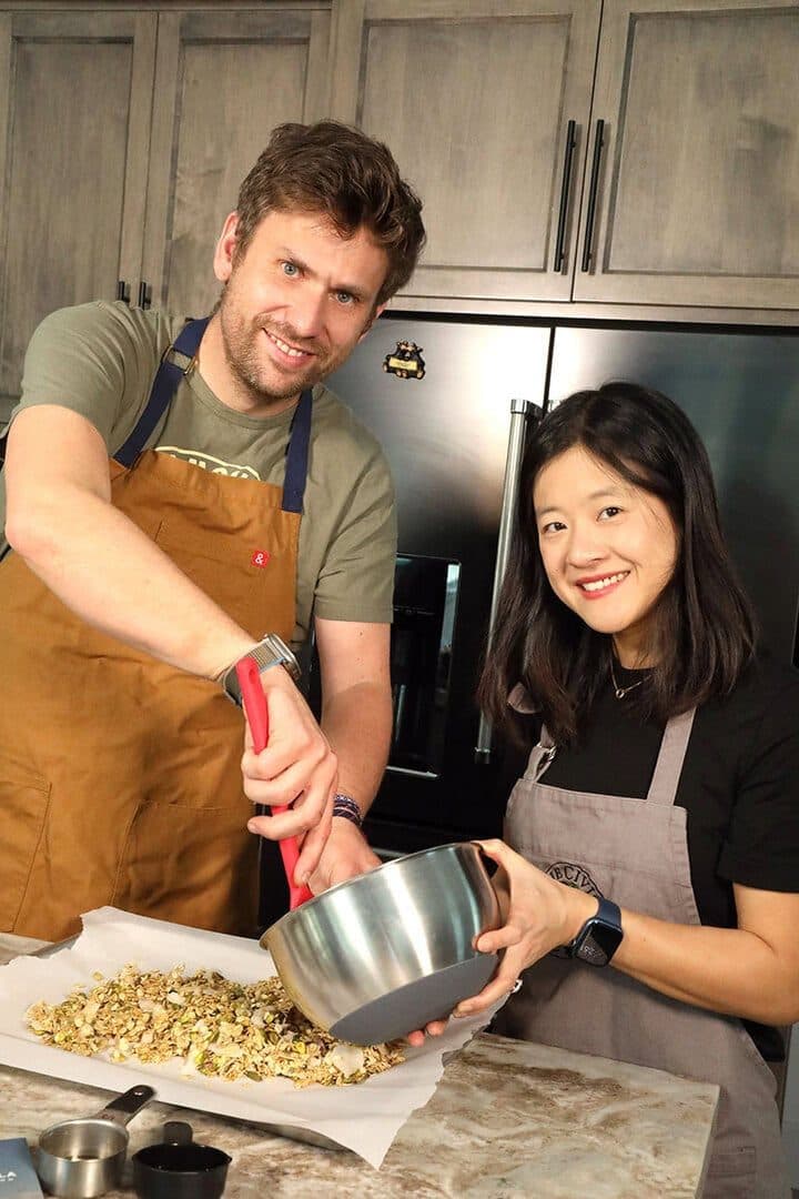 Couple in aprons mixing granola in a steel bowl in their kitchen, laughing