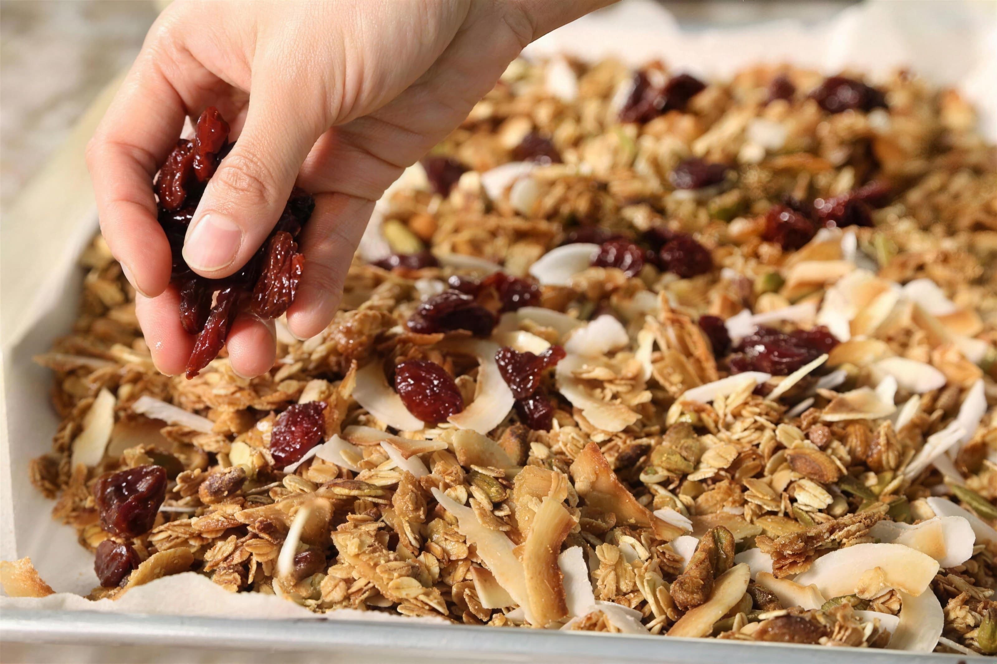Hand sprinkling dried cranberries onto a tray of golden granola with coconut flakes, close-up