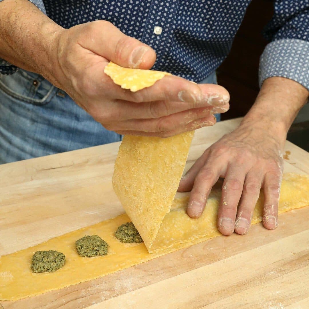 Hands folding a pasta sheet over pesto mounds to make ravioli on a wooden board