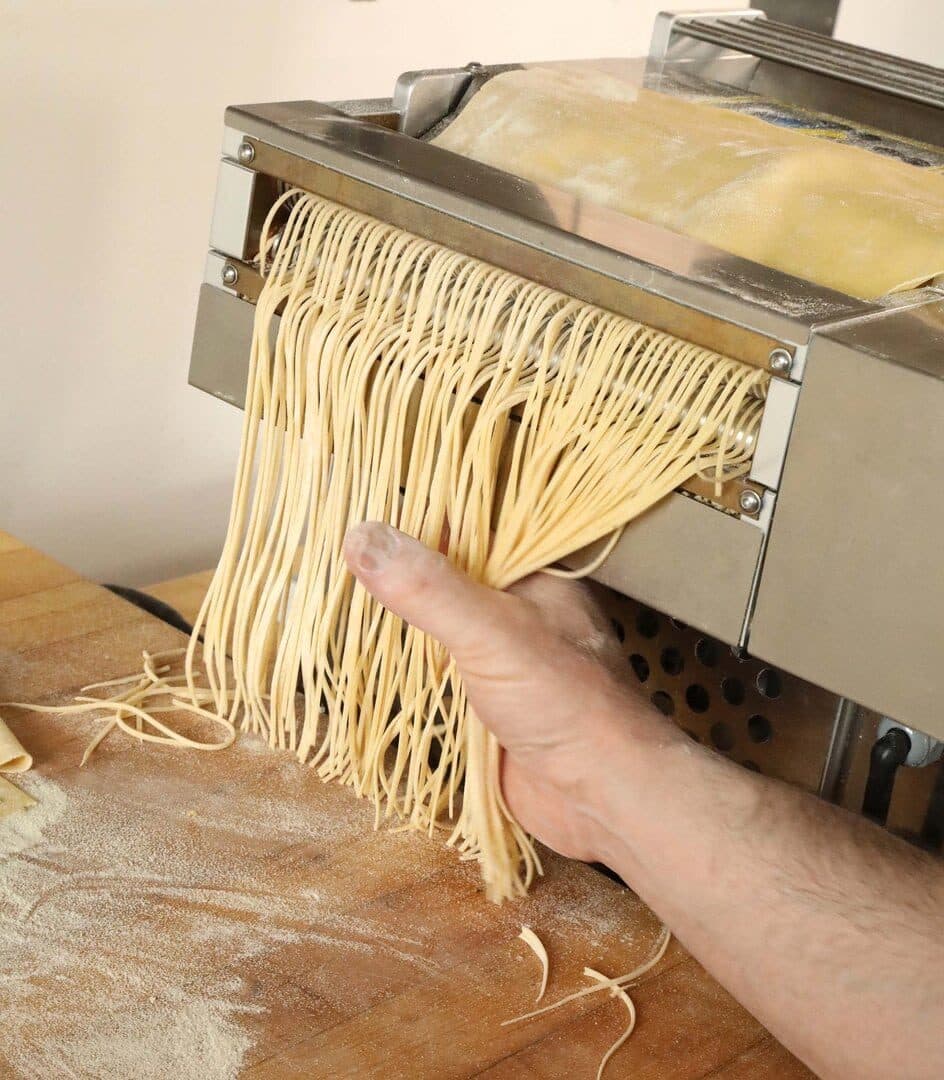 Fresh noodles extruding from a pasta machine into a hand, flour-dusted board
