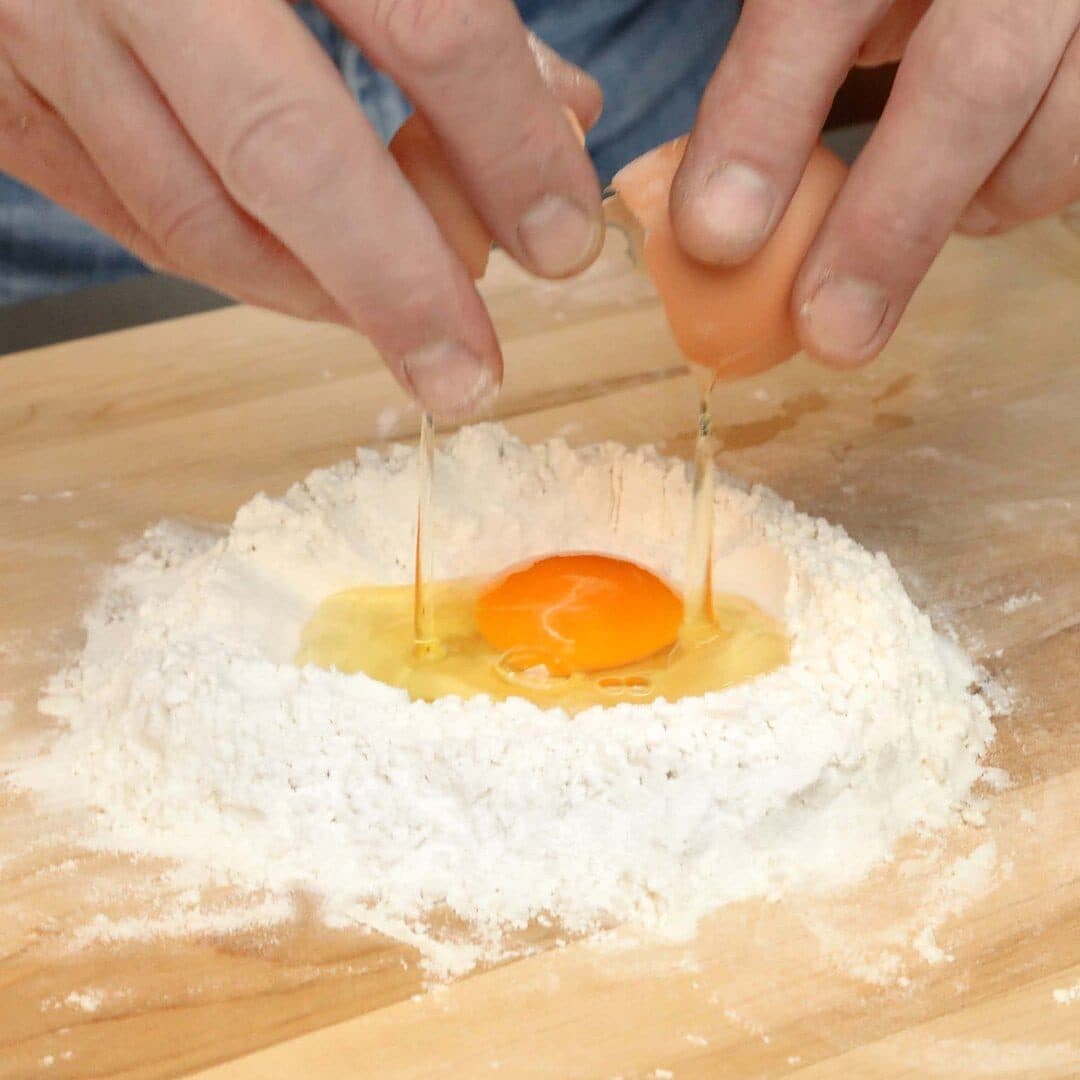 Hands cracking an egg into a well of flour, bright yolk, wooden board, close-up