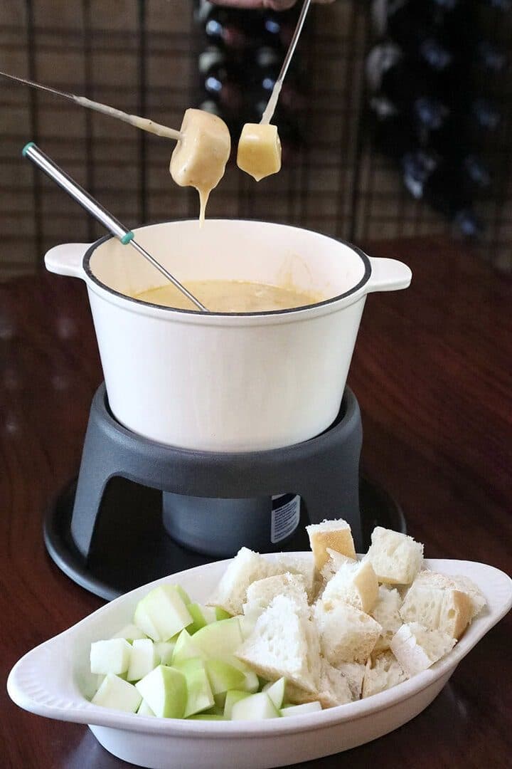 Cheese fondue pot with forks dipping bread, apples and bread cubes in a dish, dark wine-bar background