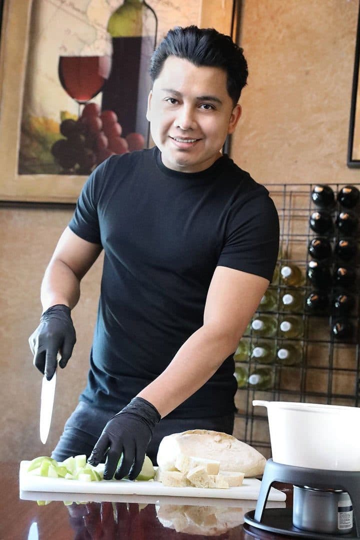 Sous chef in black gloves cutting apples beside a fondue pot, wine racks behind