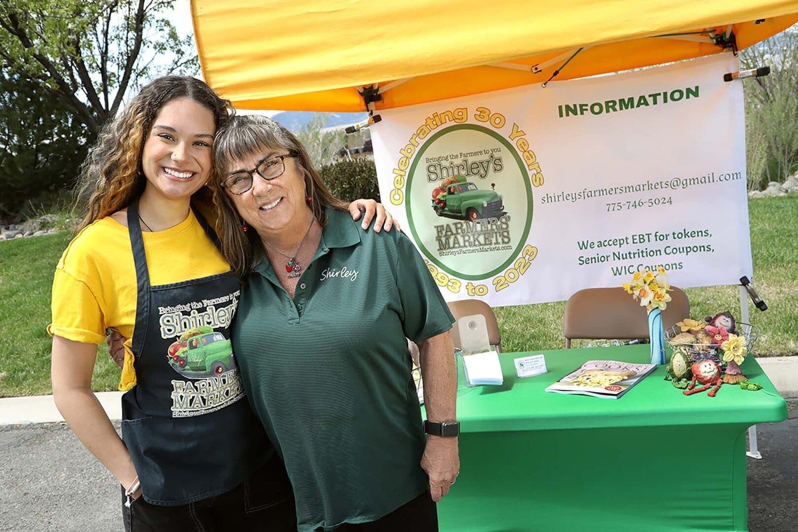 Two women smiling at a farmers' market information booth under a yellow canopy