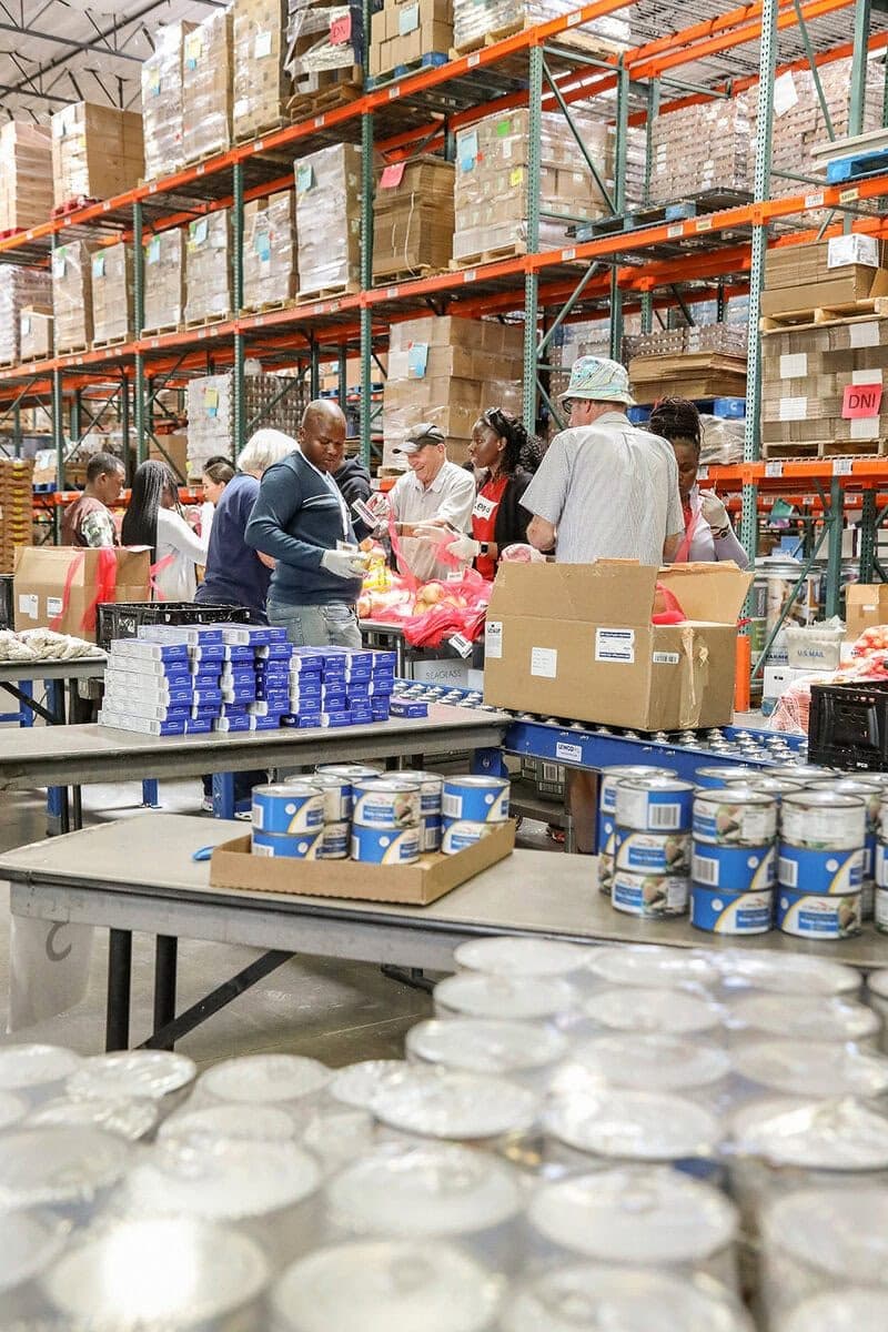 Food-bank warehouse with volunteers packing at tables, shelves of canned goods, foreground cans