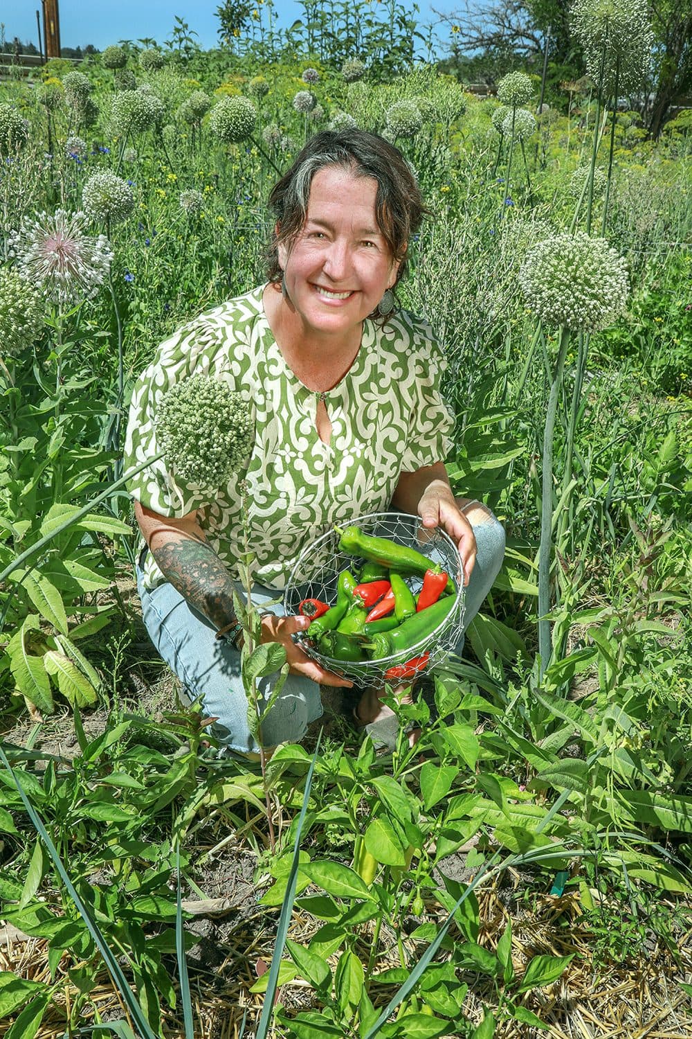 Teacher kneeling among tall allium flowers holding a basket of peppers, garden light