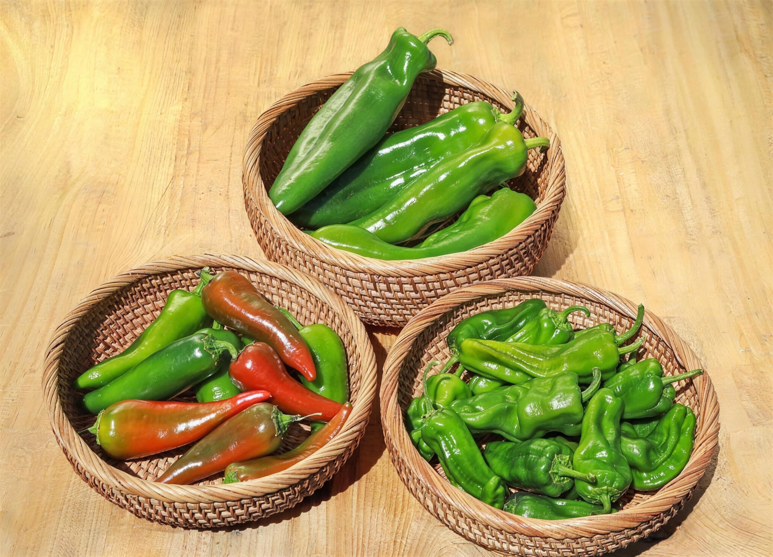 Three woven baskets of Fresno, Anaheim and Padrón peppers on a warm wood table