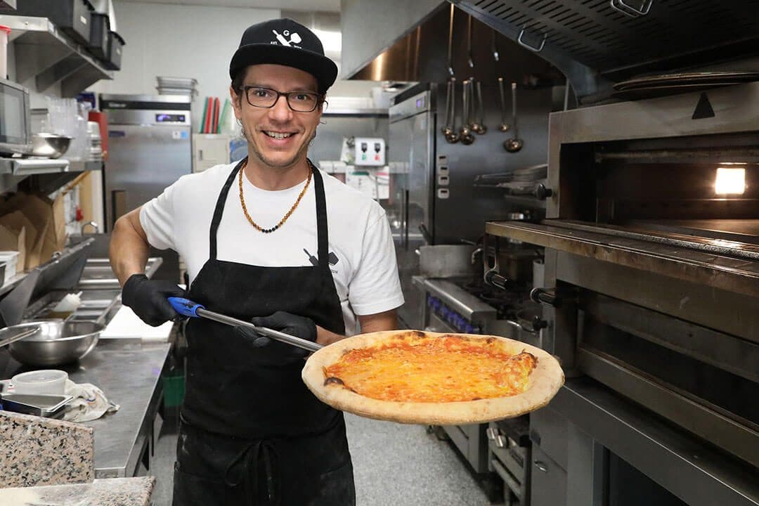Pizza chef in cap pulling a pizza from the oven on a peel, stainless kitchen