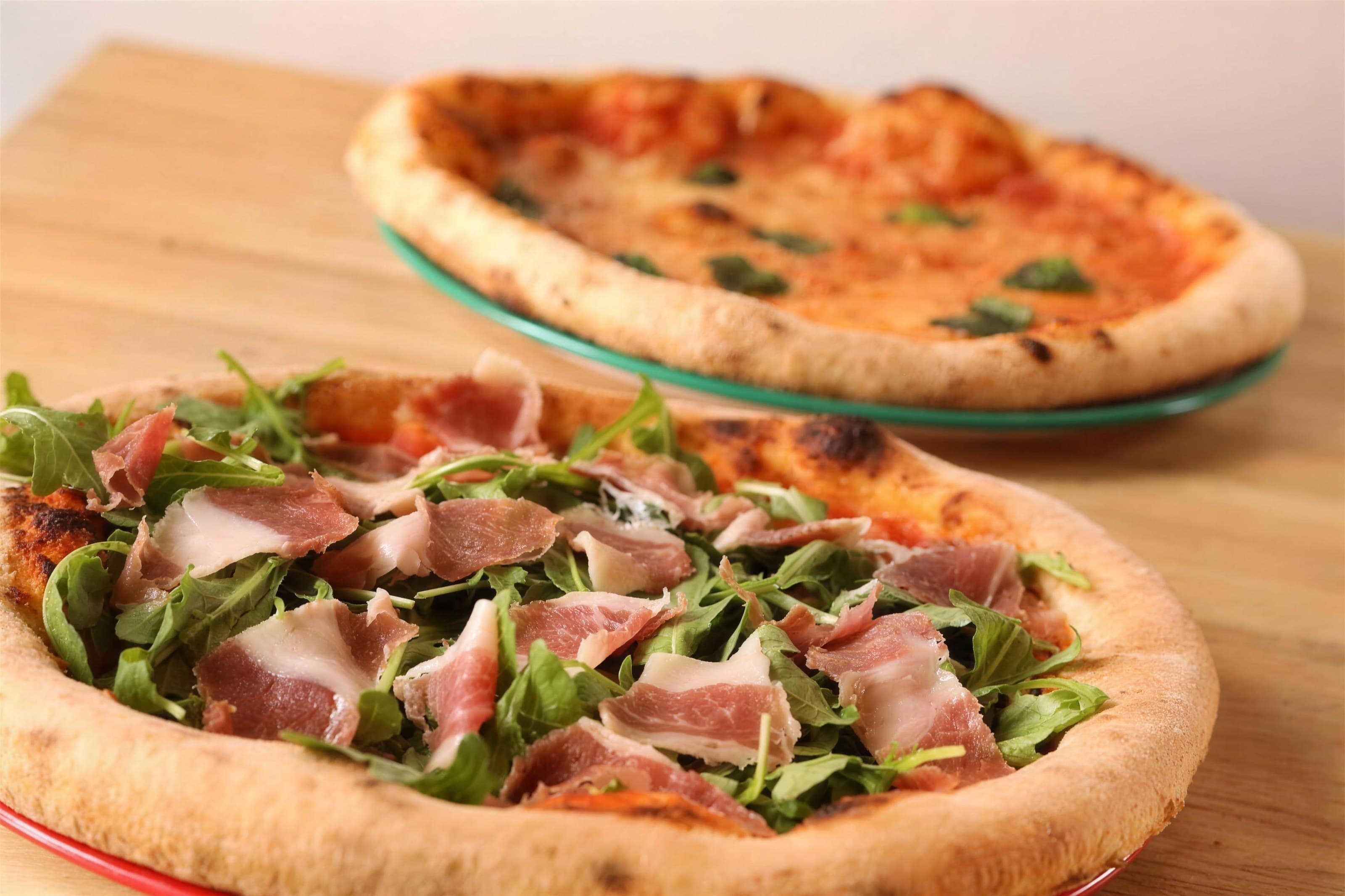 Prosciutto-arugula pizza in front of a Margherita pizza on a wood table, soft light