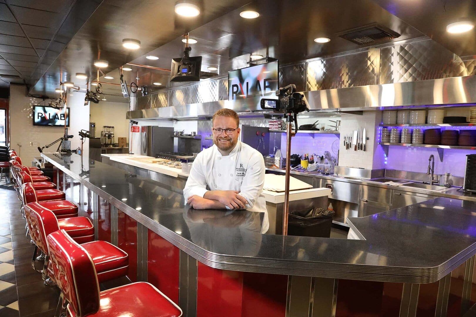 Chef Jacob Burton behind a long stainless counter in an open teaching kitchen with red stools