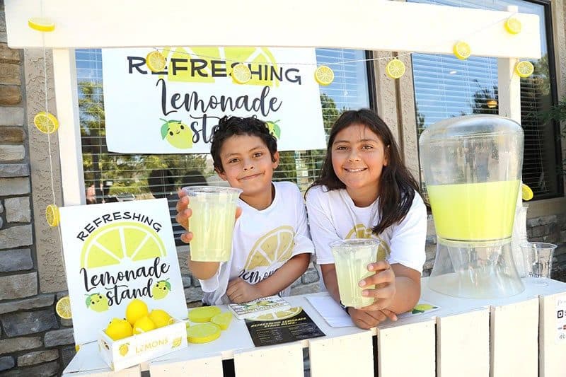 Two kids at a yellow lemonade stand holding up cups of lemonade, bright daylight