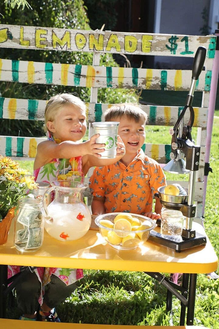 Two young kids in Hawaiian shirts at a luau-themed lemonade stand on a lawn