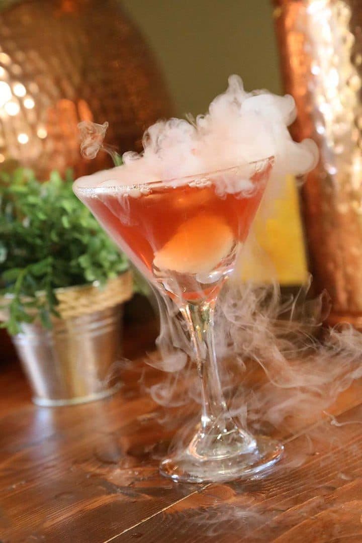 Smoking dry-ice poison apple martini on a wood bar, copper and herb backdrop, warm light