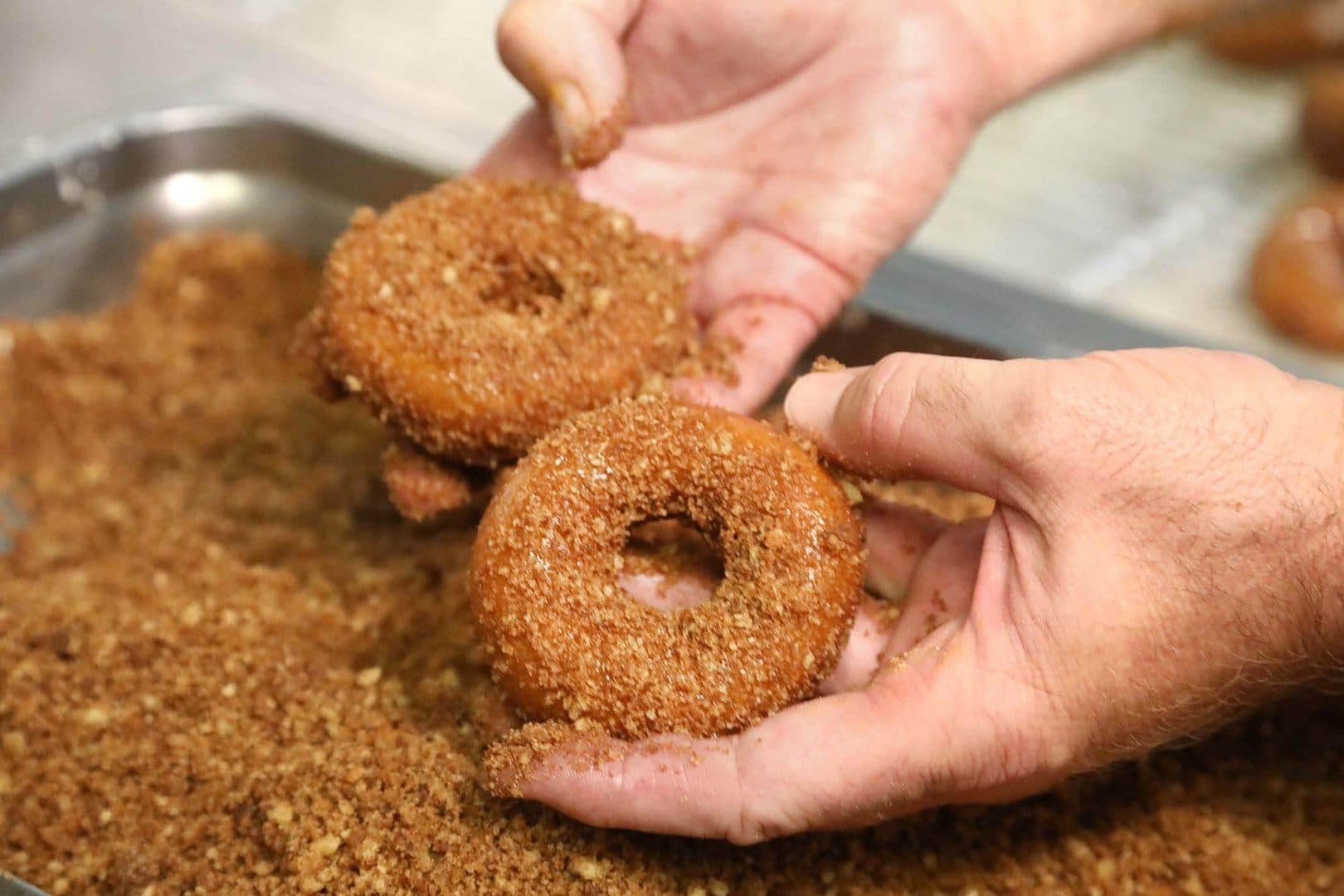 Hands pressing a doughnut into cinnamon-sugar crumble on a tray