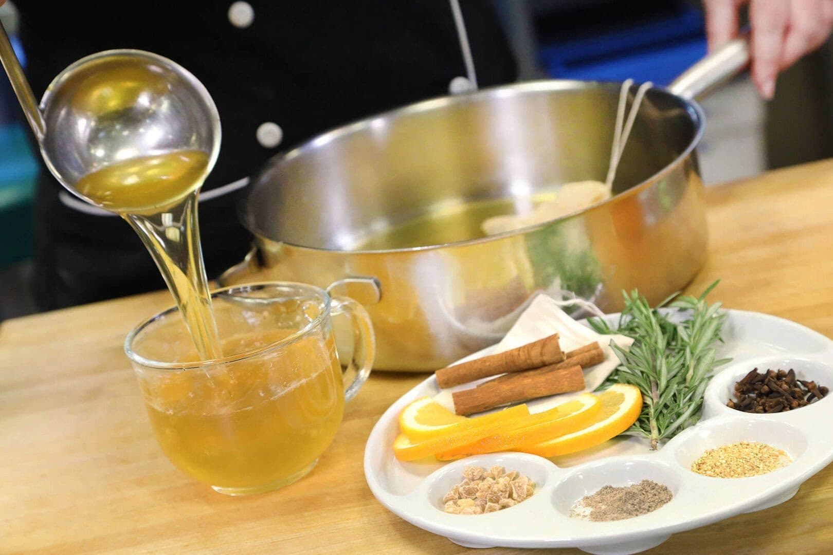Ladling mulled cider into a glass mug beside a plate of spices and orange slices