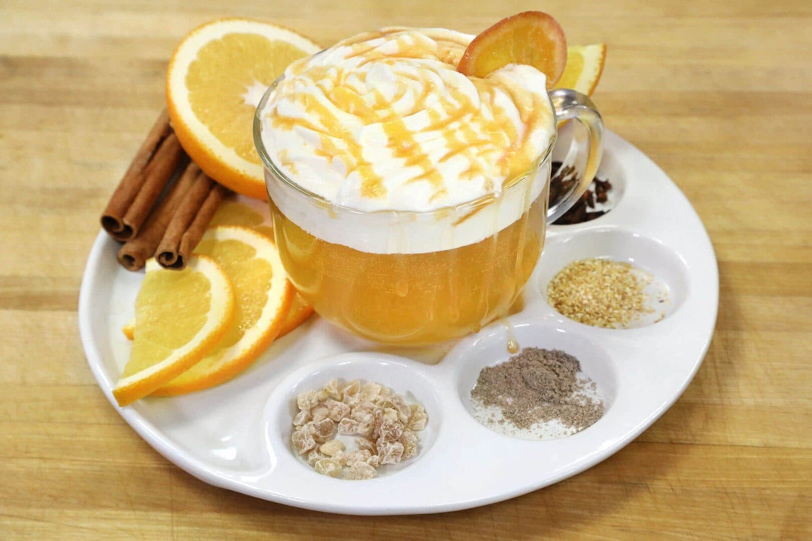 Mulled cider topped with whipped cream and caramel, oranges, cinnamon and spices on a white plate, wood counter