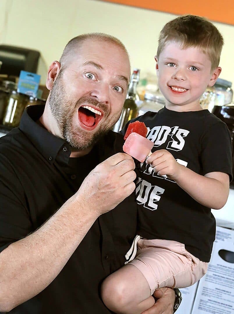 Pickle-company founder and his young son grinning with red pickle popsicles