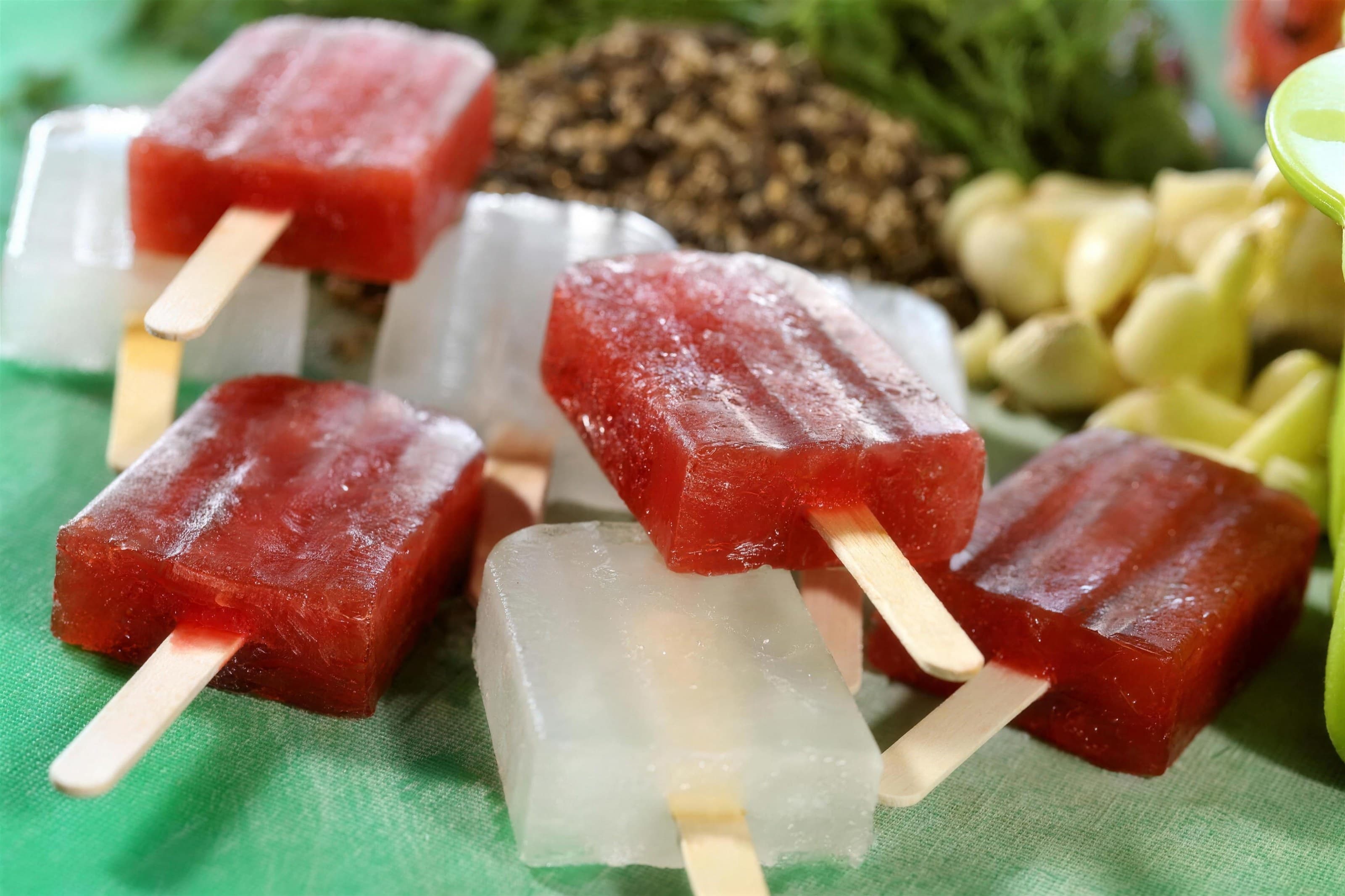 Red and clear pickle-juice popsicles on a green surface with garlic and peppercorns, bright light