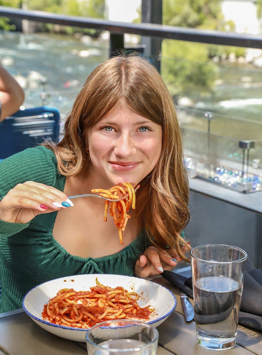 Girl twirling spaghetti marinara at a riverside restaurant table, daylight