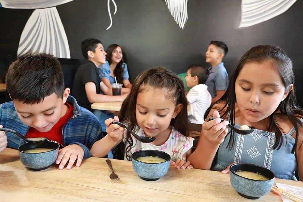 Kids eating soup at a sushi restaurant table, black-and-white wall art behind