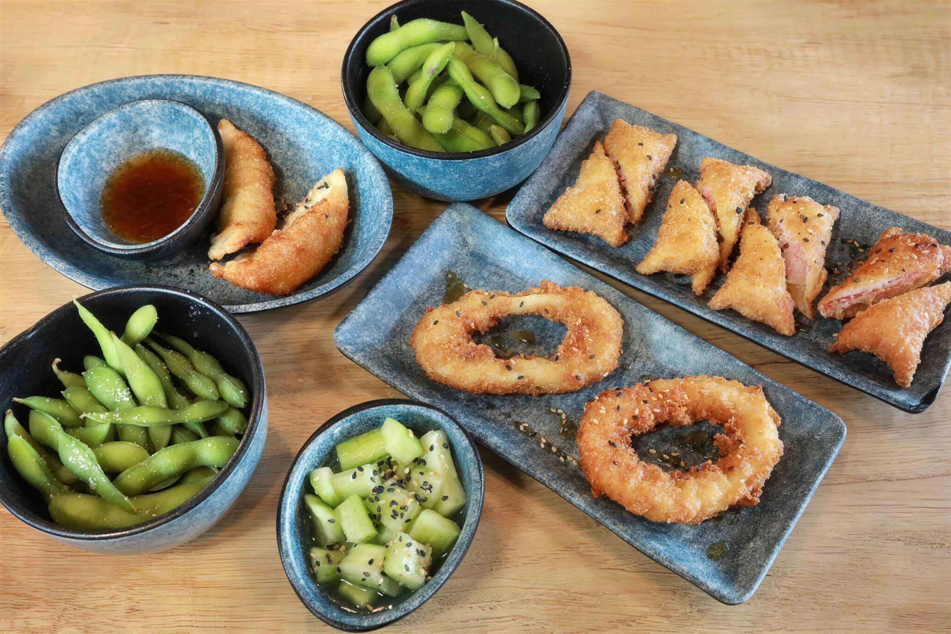 Top-down kids' sushi appetizers: edamame, gyoza, onion rings, cucumber in blue ceramic dishes