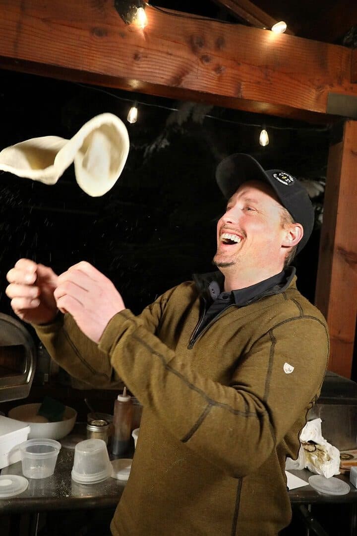 Baker tossing pizza dough overhead, laughing, warm dark restaurant