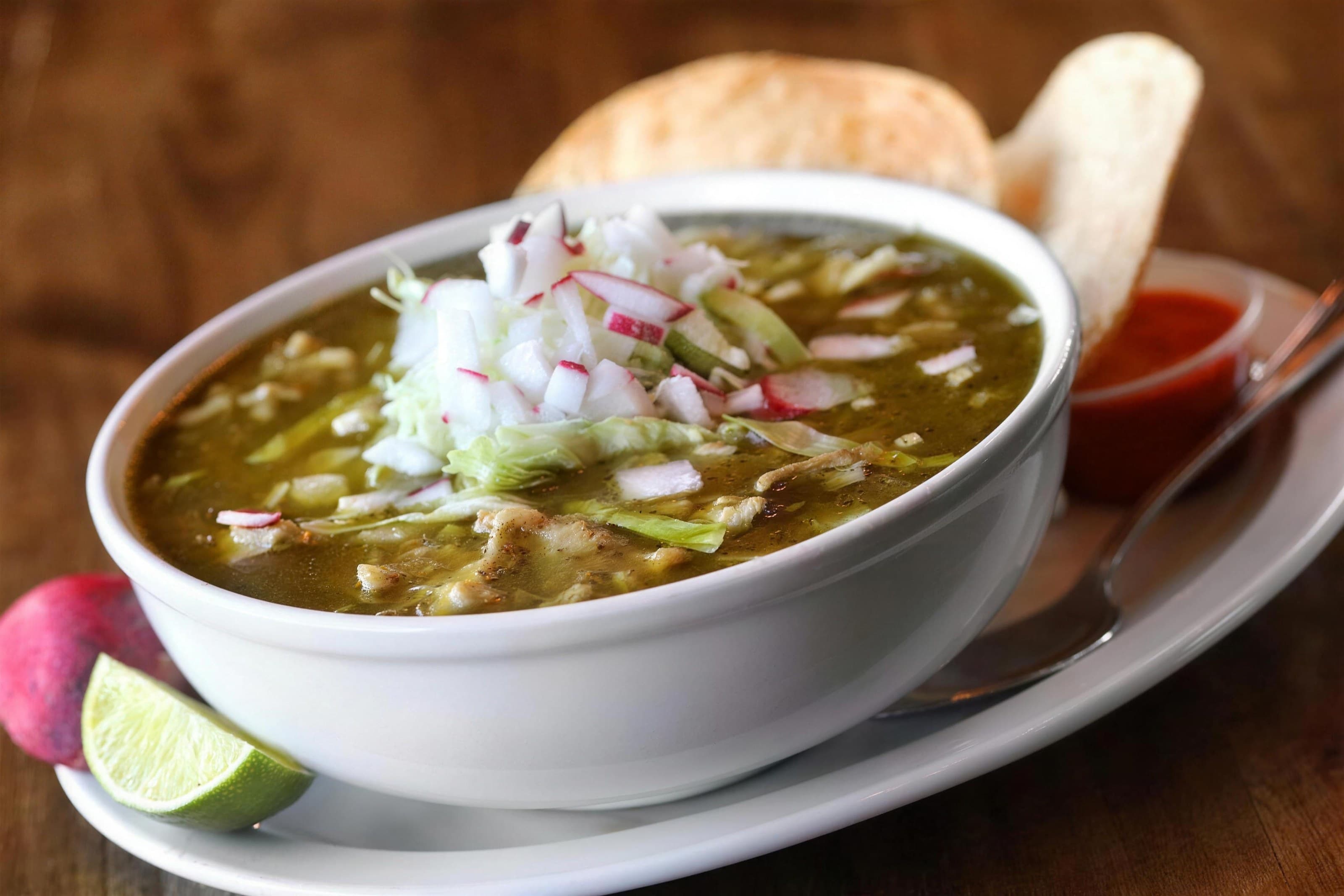 Bowl of green pozole topped with radish and onion, lime and tostadas, warm wood background