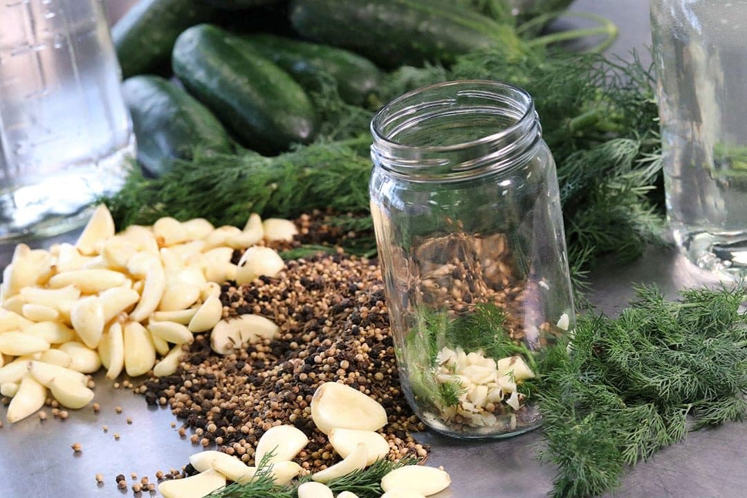 Mason jar with garlic, peppercorns and dill beside cucumbers, close-up