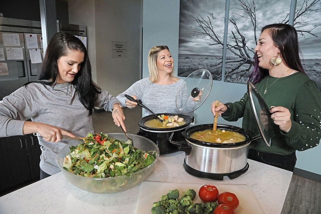 Three women serving salad and slow-cooker dishes in a modern kitchen