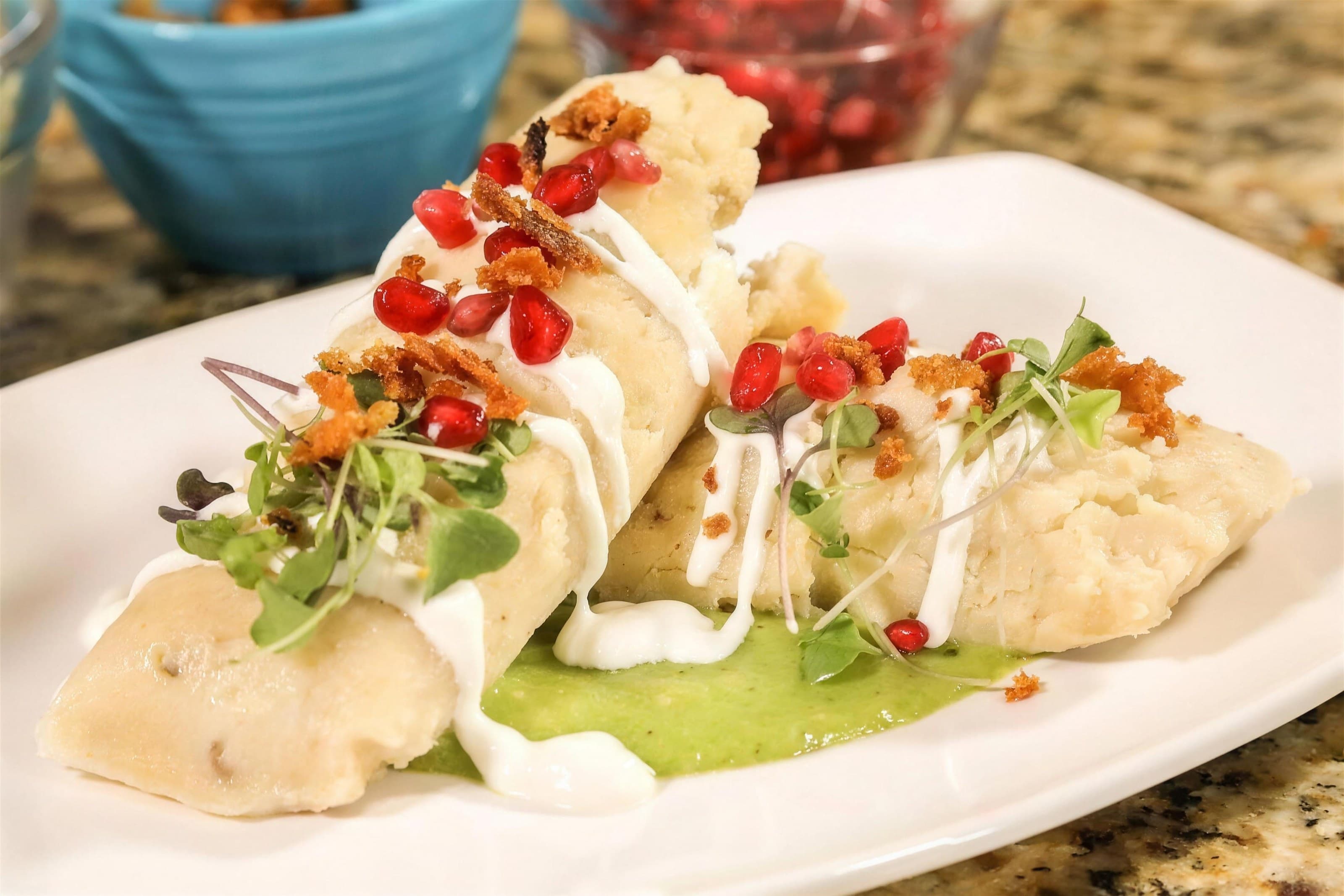 Duck tamales with pomegranate, crema and microgreens over green sauce on a white plate, granite counter