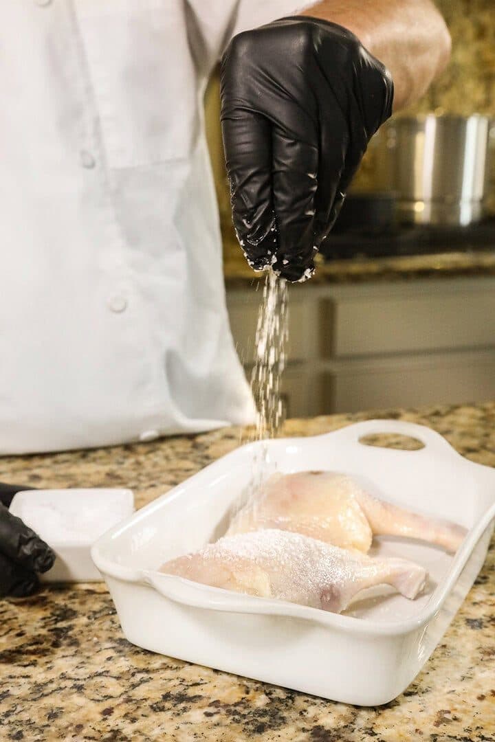 Gloved hand salting duck legs in a white dish, high-key