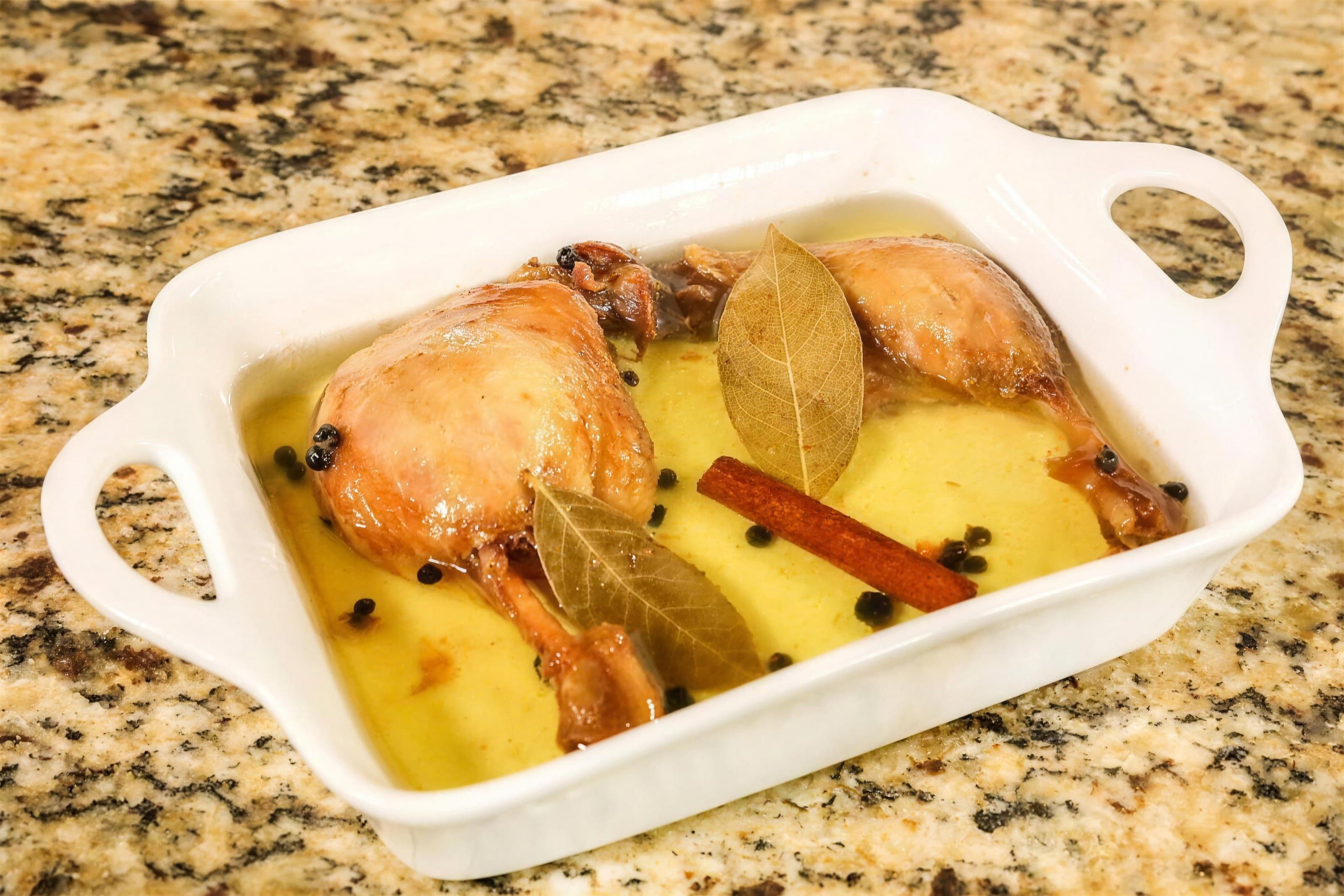 Duck leg confit in golden fat with bay leaves and cinnamon in a white baker