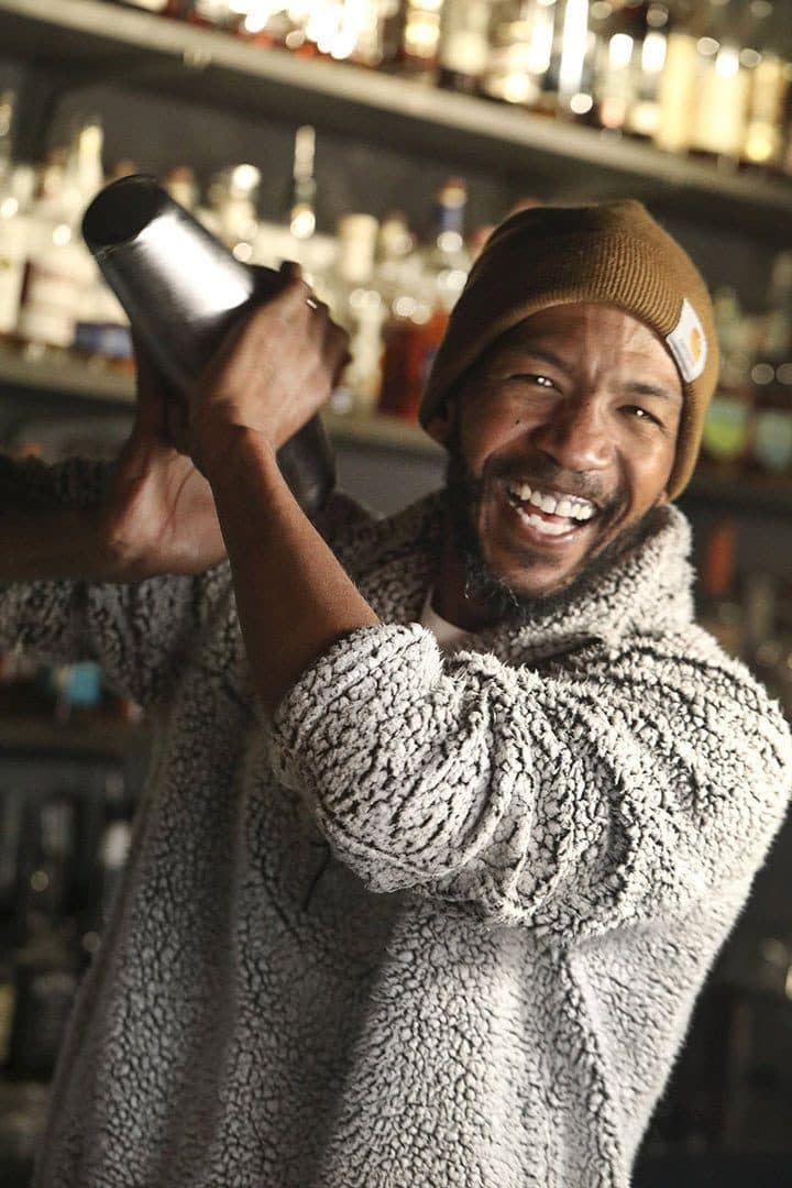 Bartender in a knit cap laughing while shaking a cocktail, back-bar bottles behind