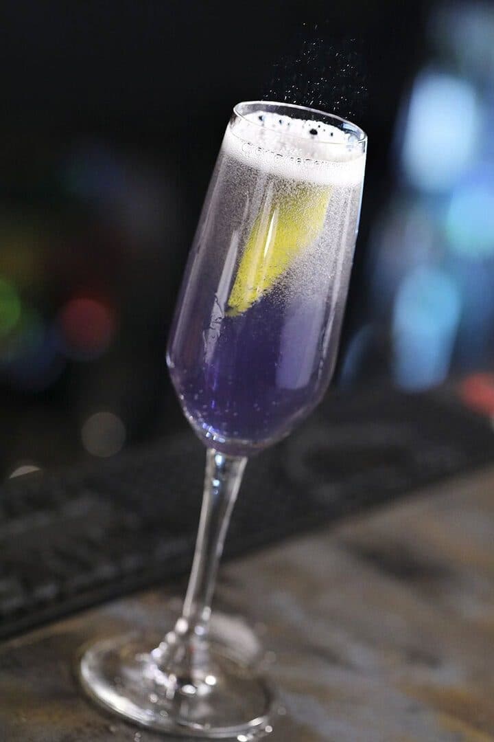 Purple Sparkling Like Royalty cocktail in a flute with lemon twist, dark bar